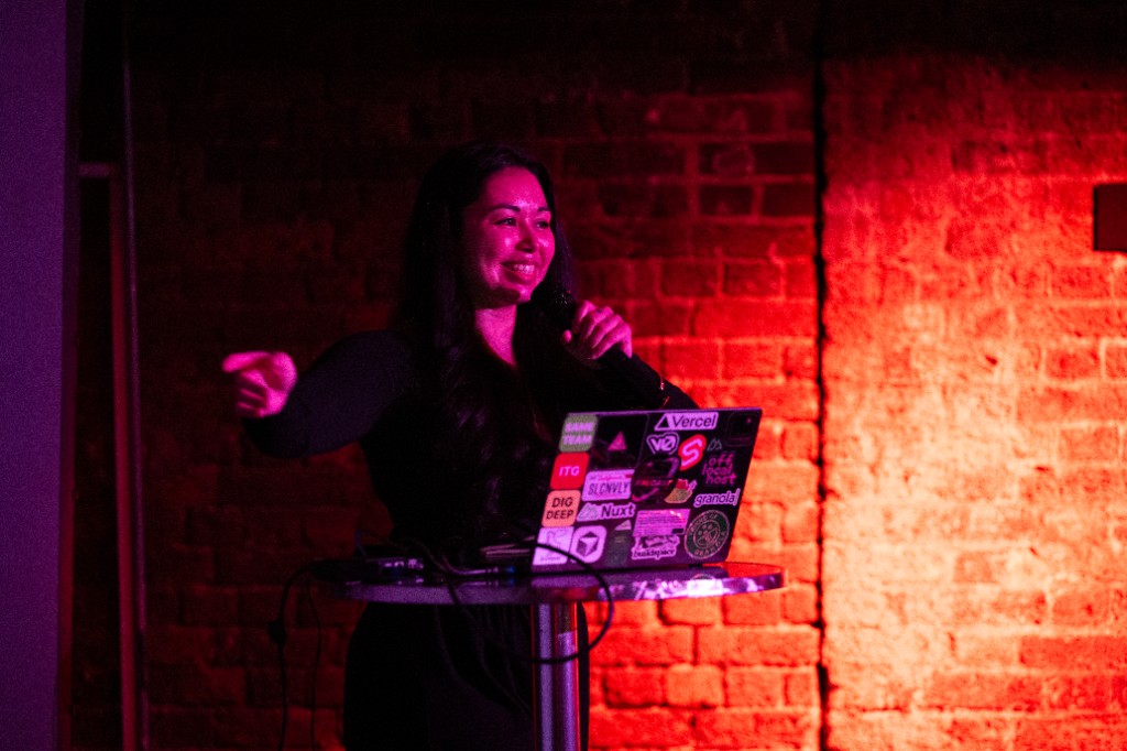 Maya speaking into a microphone at Workflows London, laptop covered in stickers on the table